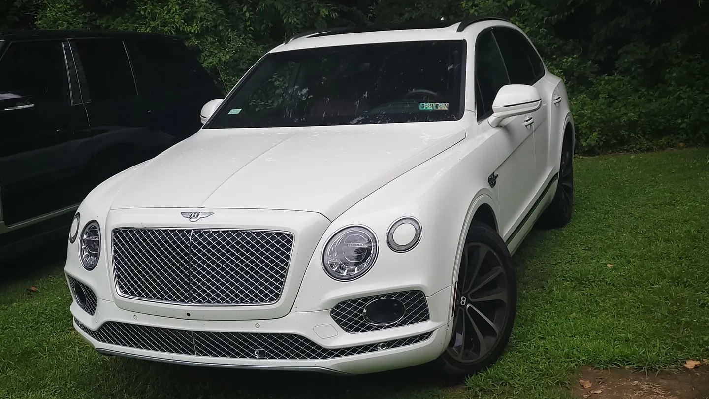 A white bentley parked in the grass near some trees.